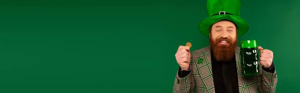 Joyeux homme au chapeau avec trèfle tenant pièce de monnaie et verre de bière isolé sur vert, bannière — Photo de stock