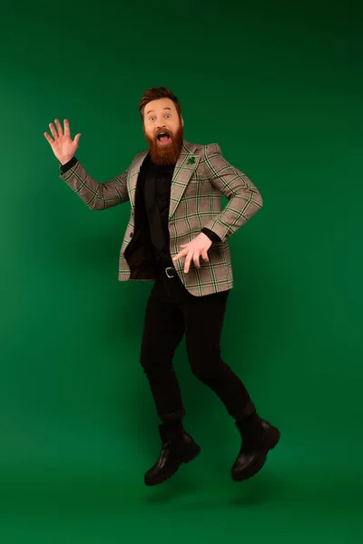 Aufgeregt bärtiger Mann in Jacke mit Kleeblatt wedelt Hand und springt auf grünem Hintergrund — Stockfoto