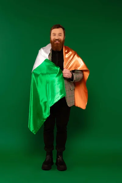 Pleine longueur d'homme barbu souriant enveloppant dans le drapeau irlandais sur fond vert — Photo de stock