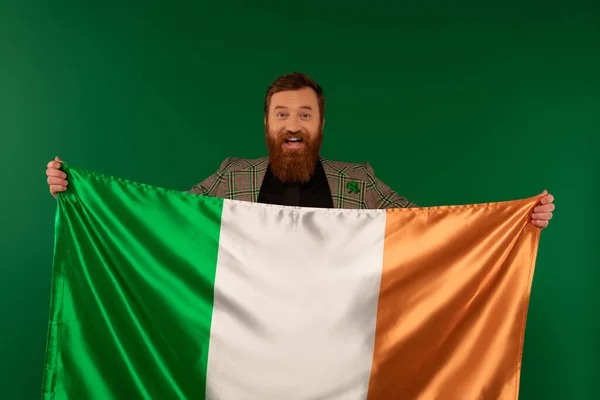 Счастливый бородатый мужчина, держащий ирландский флаг и смотрящий на камеру, изолированную на зеленом — стоковое фото