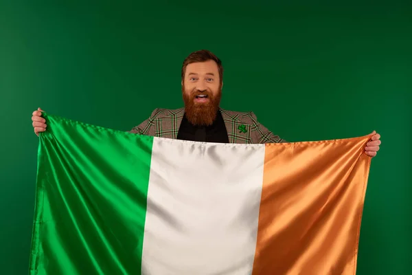 Happy bearded man holding Irish flag and looking at camera isolated on green — Stock Photo