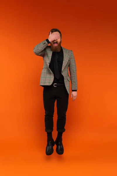 Excited bearded man covering eyes while jumping on red background - foto de stock