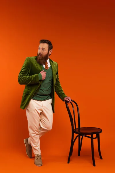 Full length of stylish man in jacket standing near chair on red background — Stock Photo
