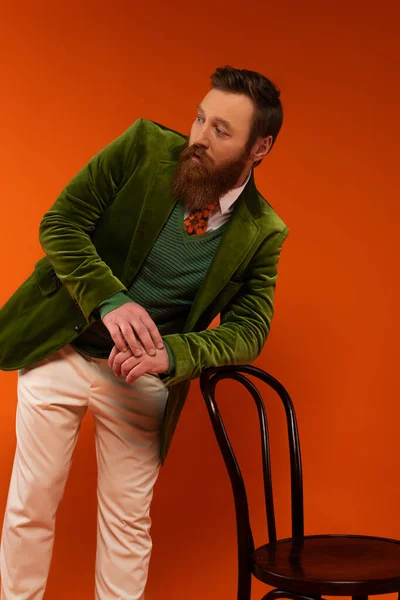 Bearded model in stylish outfit posing near chair on red background - foto de stock