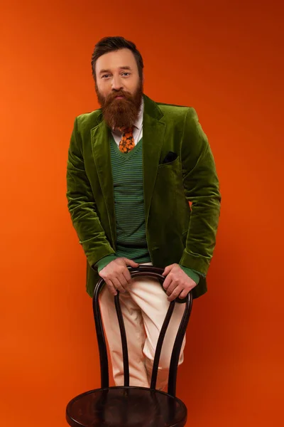 Trendy bearded man standing near chair on red background — Stock Photo