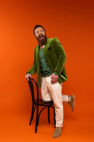 Cheerful bearded model in velvet jacket laughing near chair on red background — Stock Photo