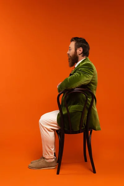 Вид збоку стильного чоловіка в оксамитовій куртці, що сидить на дерев'яному стільці на червоному тлі — стокове фото
