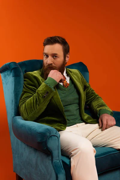Trendy bearded model looking at camera while sitting on armchair on red background - foto de stock