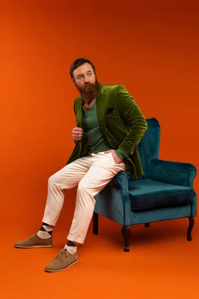 Стильный бородатый мужчина позирует возле бархатного кресла на красном фоне — стоковое фото