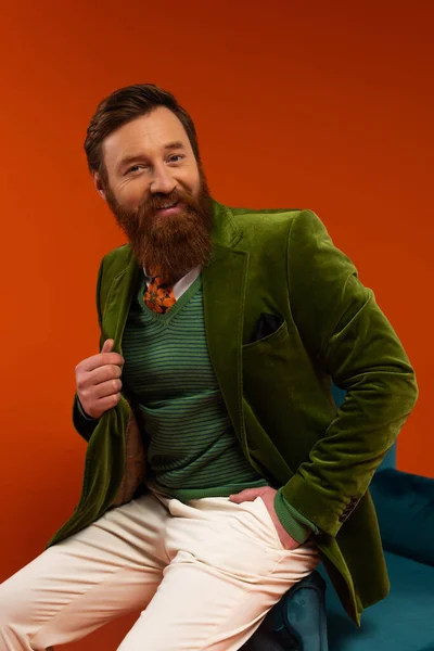 Smiling bearded man in stylish outfit sitting on armchair on red background - foto de stock