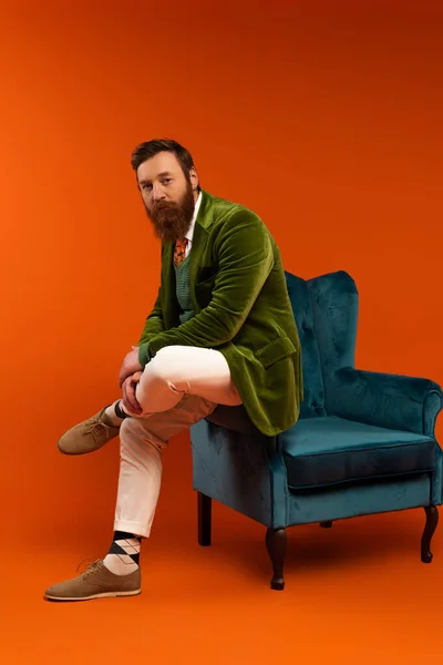 Trendy bearded model in stylish clothes sitting on blue armchair on red background — Photo de stock