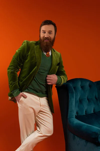 Stylish bearded man looking at camera while posing near armchair on red background — Stock Photo