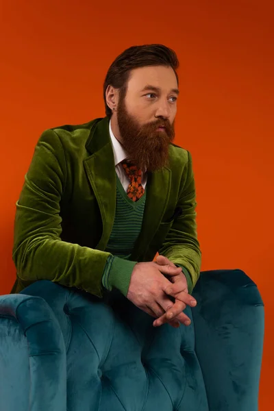 Trendy bearded model in velvet blazer standing near armchair on red background — Stock Photo