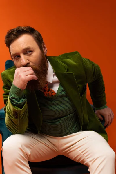 Fashionable bearded man in jacket sitting on armchair on red background - foto de stock