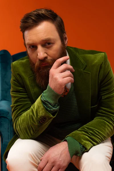 Trendy bearded model in velour jacket sitting on armchair on red background - foto de stock