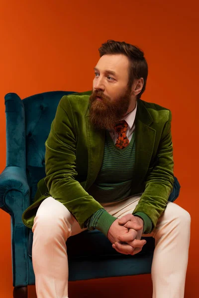 Модный бородатый мужчина сидит на синем бархатном кресле на красном фоне — стоковое фото