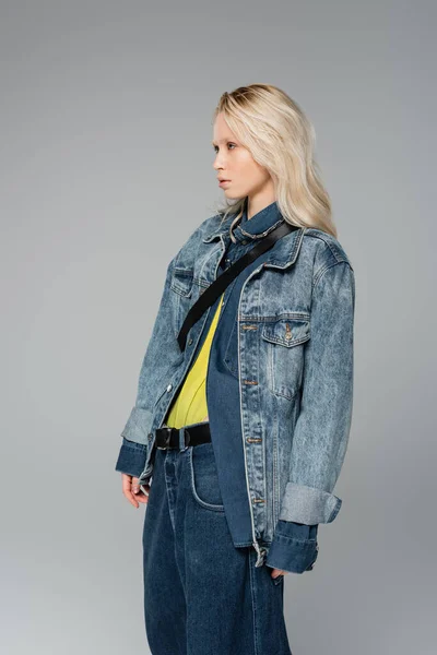 Junges blondes Model im stylischen Jeans-Outfit schaut weg, während sie isoliert auf grau posiert — Stockfoto