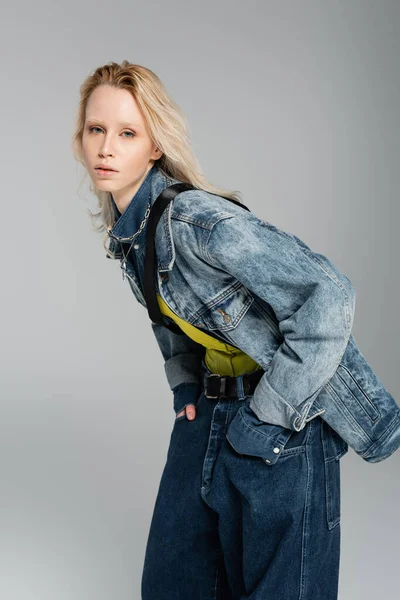 Junges blondes Model in stylischem Jeans-Outfit blickt in die Kamera, während sie mit Händen in Taschen posiert, die isoliert auf grau sind — Stockfoto