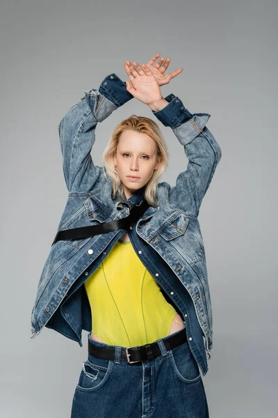 Young blonde woman in stylish denim outfit looking at camera while posing with hands above head isolated on grey — Fotografia de Stock
