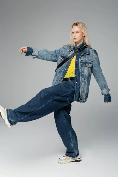 In voller Länge junge blonde Frau im trendigen Jeans-Outfit posiert beim Gehen auf grau — Stockfoto