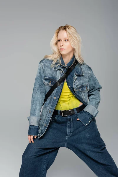 Молодая блондинка в стильной джинсовой одежде позирует и смотрит в сторону изолированной на сером — стоковое фото