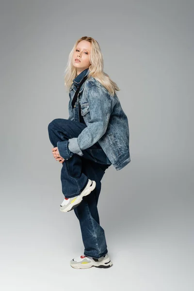 Pleine longueur de jeune femme blonde en tenue de denim élégant et baskets posant tout en se tenant debout sur une jambe sur gris — Photo de stock
