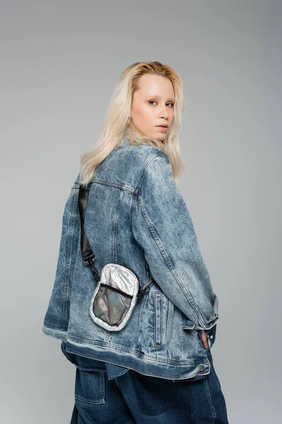 Молодая блондинка в стильной джинсовой куртке с ременной сумкой позирует изолированно на сером — стоковое фото
