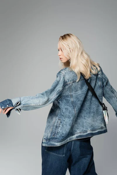 Junge blonde Frau in stylischer Jeansjacke mit Gürteltasche posiert isoliert auf grau — Stockfoto