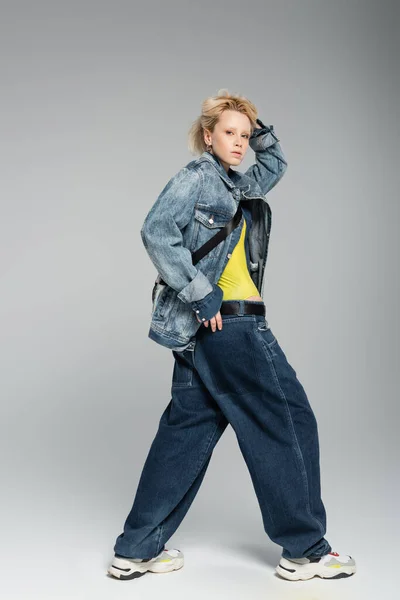 Full length of young blonde model in stylish denim outfit and trendy sneakers posing on grey — Stock Photo