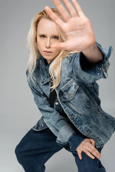Young blonde model in trendy denim outfit looking at camera while posing with outstretched hand isolated on grey — Photo de stock