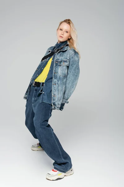 Full length of blonde model in stylish denim outfit and trendy sneakers posing on grey — Photo de stock