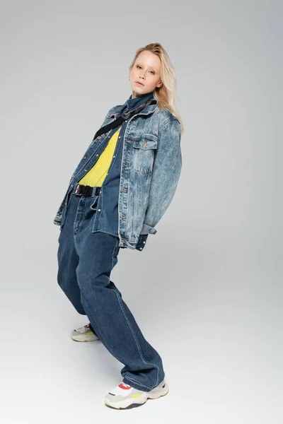 Full length of blonde model in stylish denim outfit and trendy sneakers posing on grey — Stock Photo