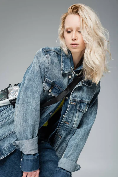 Young blonde model in stylish denim jacket and belt bag posing isolated on grey — Stock Photo
