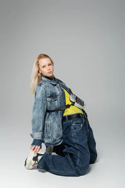 Full length of young blonde woman in blue denim outfit and trendy sneakers posing on grey — Photo de stock