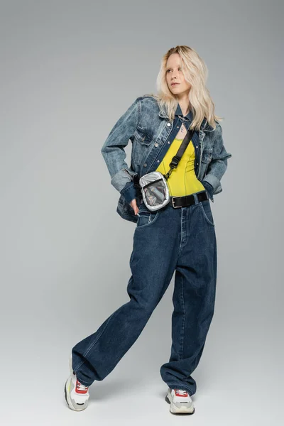 Longitud completa de la mujer rubia en traje de mezclilla azul y zapatillas de deporte de moda de pie en gris — Stock Photo