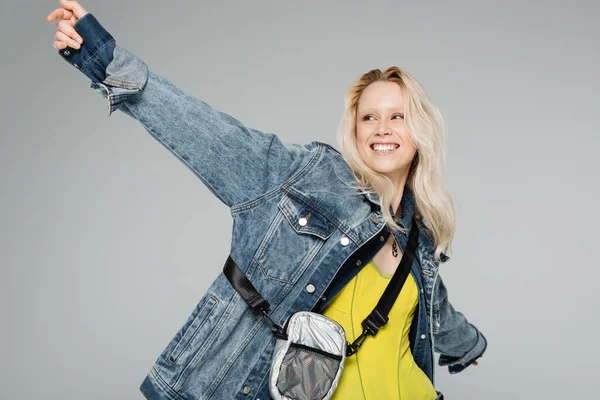 Glückliche blonde Frau in stylischer Jeansjacke mit Gürteltasche stehend mit ausgestreckten Händen isoliert auf grau — Stockfoto