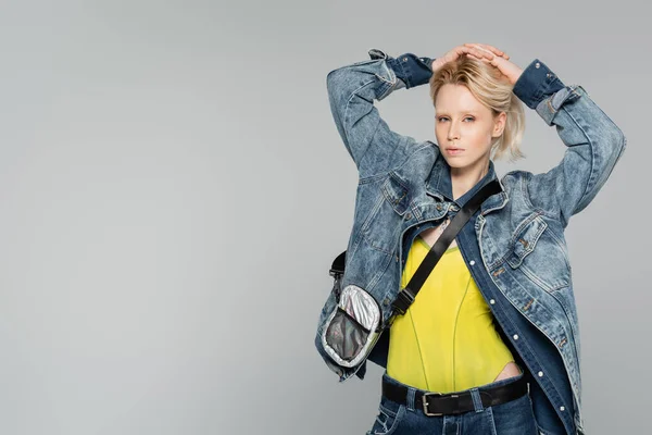 Joven mujer rubia en elegante chaqueta de mezclilla posando con las manos en la cabeza aislado en gris — Stock Photo