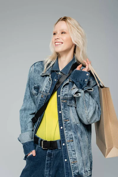 Mujer joven positiva en traje de mezclilla elegante sosteniendo bolsas de compras mientras posando con la mano en el bolsillo aislado en gris - foto de stock
