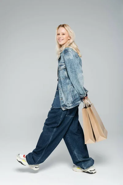 Full length of happy young woman in stylish denim outfit holding shopping bags and walking on grey — Photo de stock
