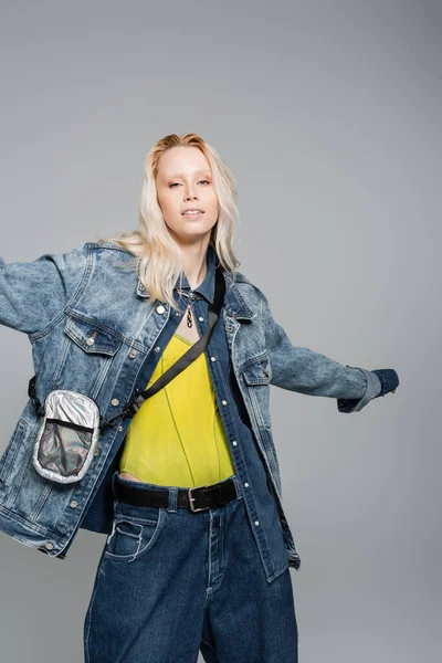 Young blonde woman in denim blue jacket and belt bag posing with outstretched hand isolated on grey — Stock Photo