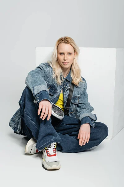 Full length of blonde woman in blue denim outfit and trendy sneakers sitting near white cube on grey — Stock Photo