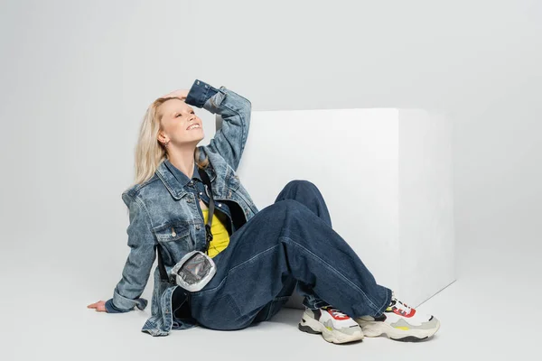 Longitud completa de la mujer positiva en traje de mezclilla azul y zapatillas de deporte de moda sentado cerca del cubo blanco en gris - foto de stock