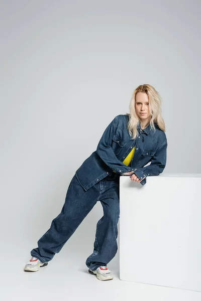 Junges blondes Model in blauem Jeans-Outfit und trendigen Turnschuhen, angelehnt an weißen Würfel auf grau — Stockfoto