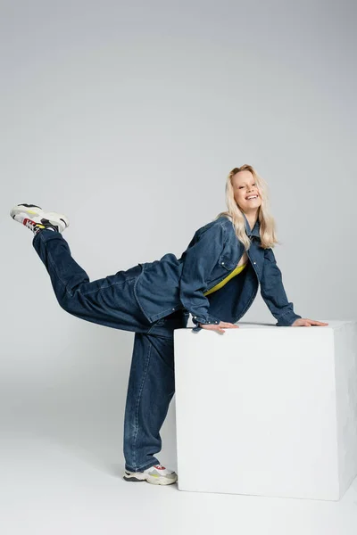 Full length of positive woman in denim outfit and trendy sneakers posing with raised leg near white cube on grey - foto de stock