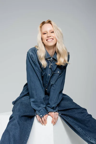 Happy woman in denim jacket and trendy jeans sitting on white cube isolated on grey — Stock Photo