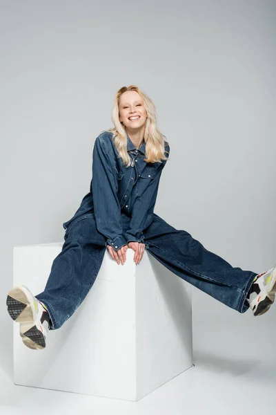 Full length of happy woman in denim outfit and trendy sneakers sitting on white cube on grey — Stock Photo