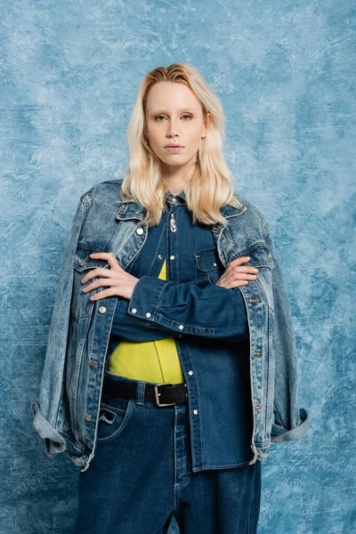 Blonde model in denim jacket posing with crossed arms near blue textured background — Stock Photo