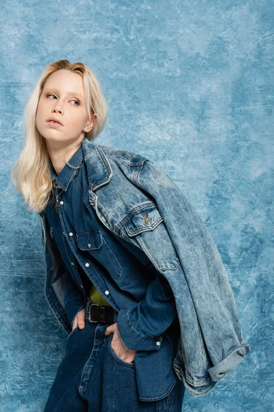 Blonde woman in denim jacket posing with hands in pockets near blue textured background — Stock Photo