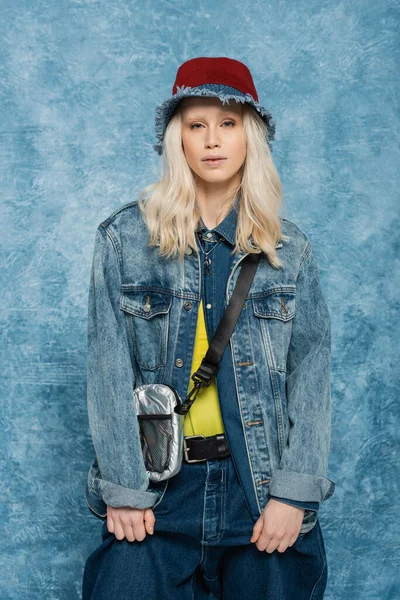 Блондинка в джинсовой куртке и в панамской шляпе позирует возле синего текстурированного фона — стоковое фото