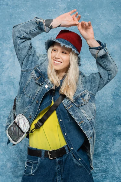 Cheerful woman in denim jacket and panama hat posing with raised hands near blue textured background — Stock Photo